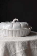 A glossy white ceramic pumpkin-shaped lidded dish with a ribbed base, set on a draped white linen cloth against a dark black background.