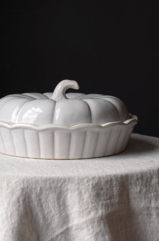 A glossy white ceramic pumpkin-shaped lidded dish with a ribbed base, set on a draped white linen cloth against a dark black background.