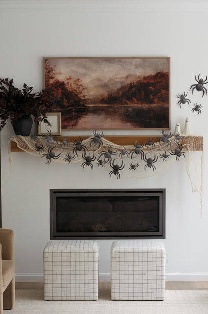 A styled interior scene showing a white wall with a framed dark rectangular mirror or picture, a vertical abstract painting in brown, black, and beige tones, a decorative mantel with a spider garland, and two upholstered ottomans on the left.