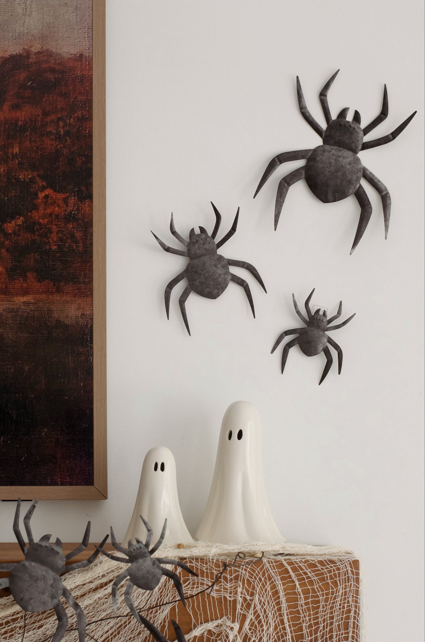 Halloween decoration set featuring black plastic toy spiders and two white plastic ghost decorations arranged on a white wall next to a wooden board with fake cobwebs.