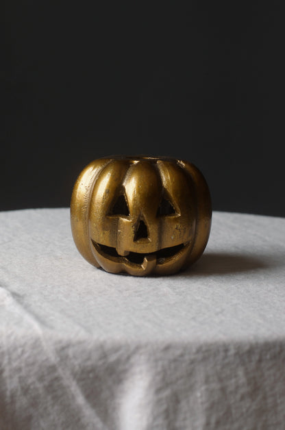 A small round gold-toned button with a carved jack-o'-lantern face, shown on a white fabric-covered display against a dark gray background.