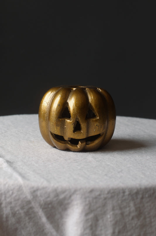 A small round gold-toned button with a carved jack-o'-lantern face, shown on a white fabric-covered display against a dark gray background.
