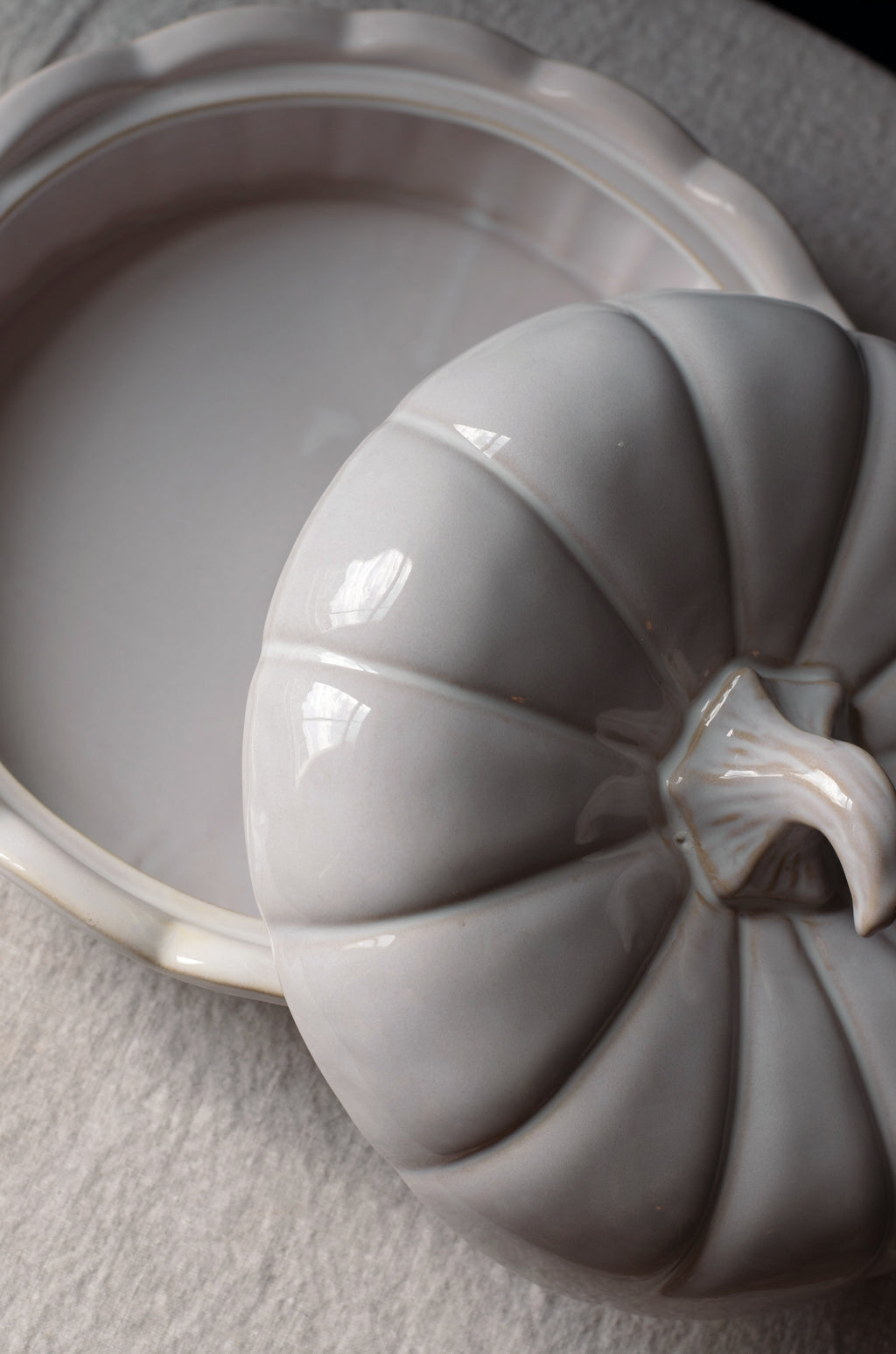 A close-up of a glossy ceramic pumpkin-shaped lidded dish resting beside an open matching pumpkin-shaped dish on a light textured surface.