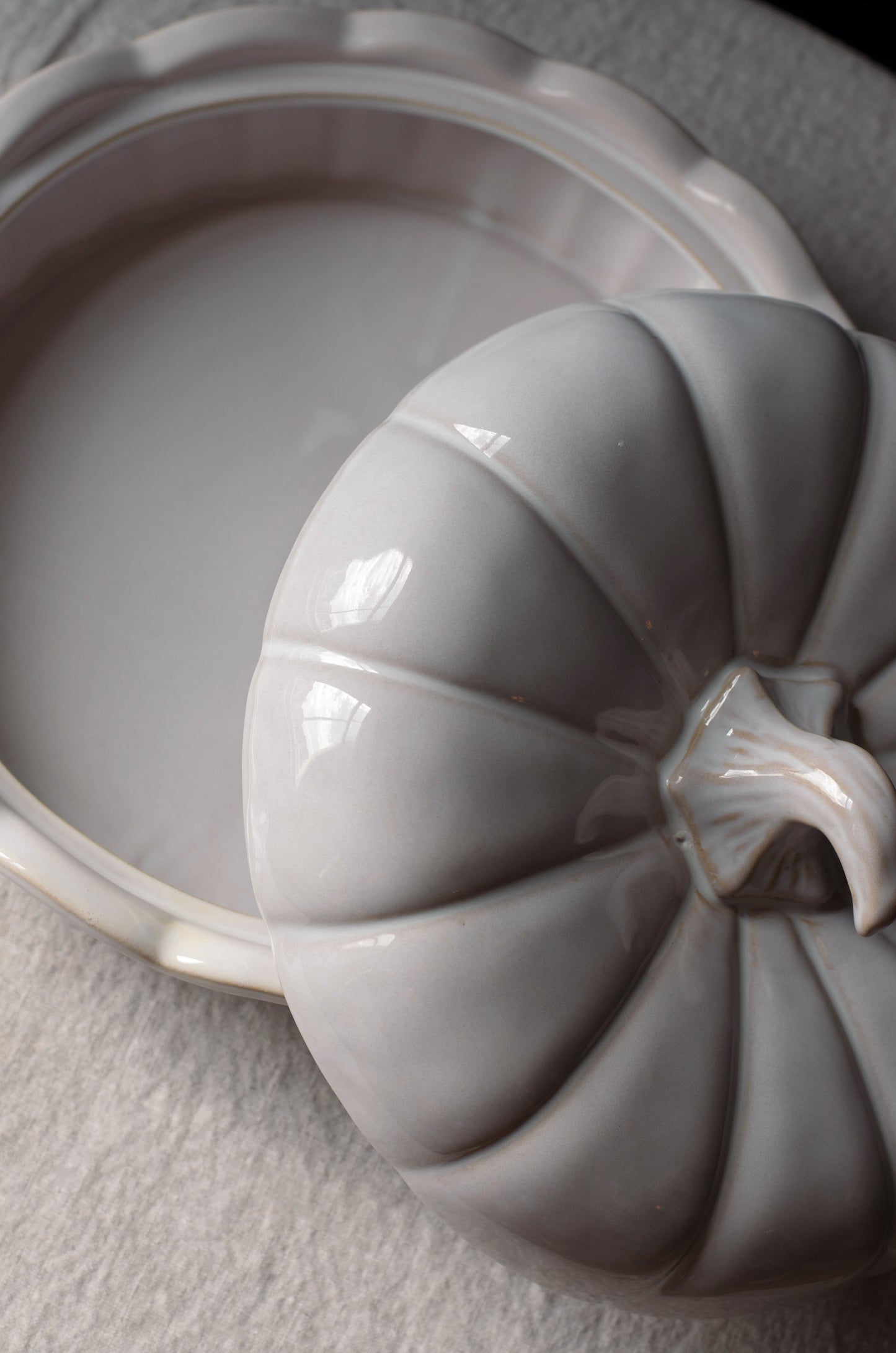 A close-up of a glossy ceramic pumpkin-shaped lidded dish resting beside an open matching pumpkin-shaped dish on a light textured surface.