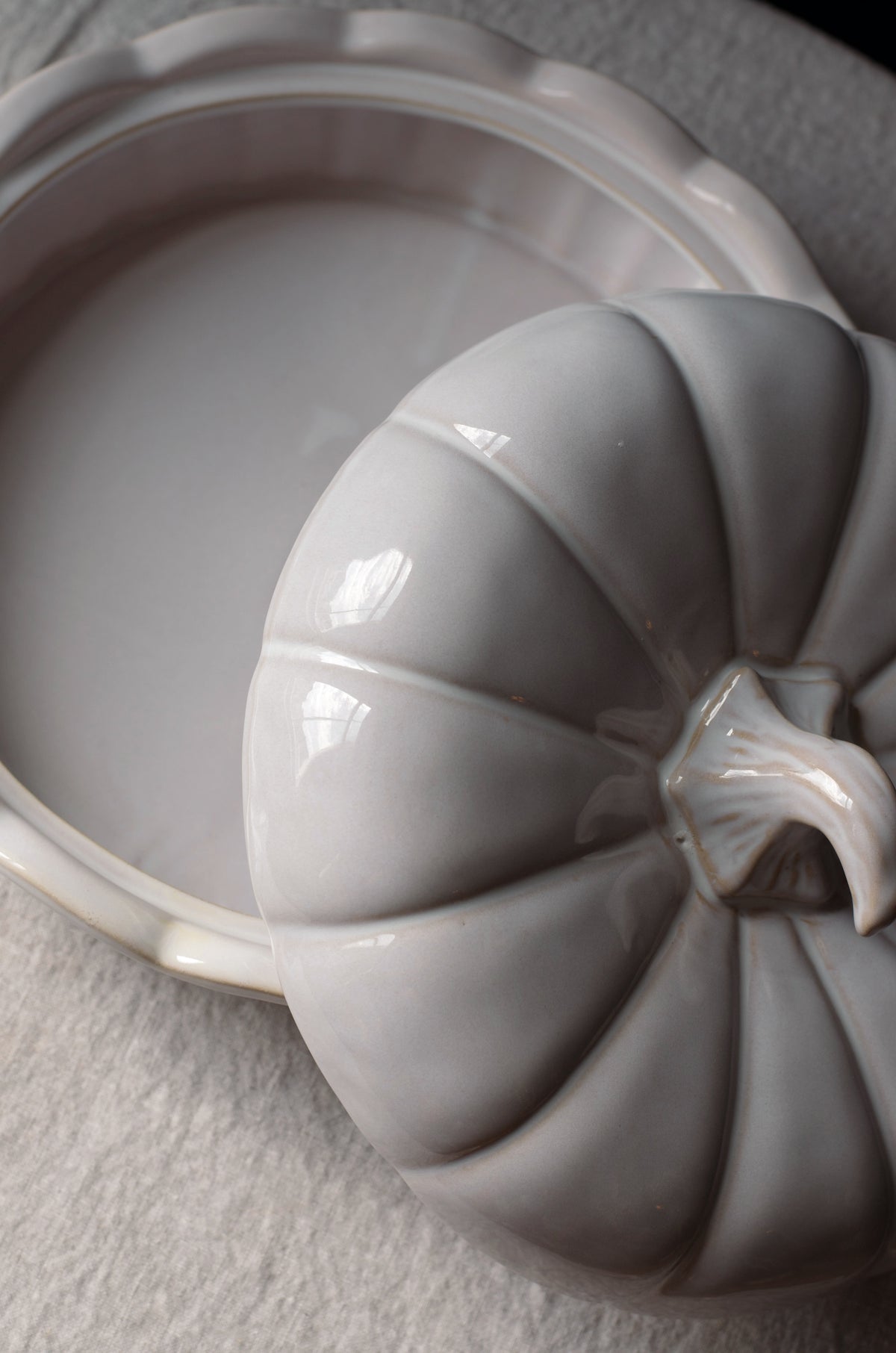 A close-up of a glossy ceramic pumpkin-shaped lidded dish resting beside an open matching pumpkin-shaped dish on a light textured surface.