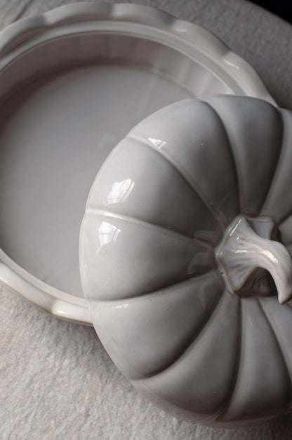 A close-up of a glossy ceramic pumpkin-shaped lidded dish resting beside an open matching pumpkin-shaped dish on a light textured surface.