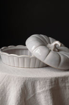 A glossy white ceramic lidded dish or container shaped like a ribbed pumpkin, shown with its lid resting partly open on a draped white fabric surface against a dark background.
