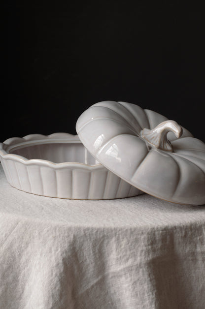 A glossy white ceramic lidded dish or container shaped like a ribbed pumpkin, shown with its lid resting partly open on a draped white fabric surface against a dark background.