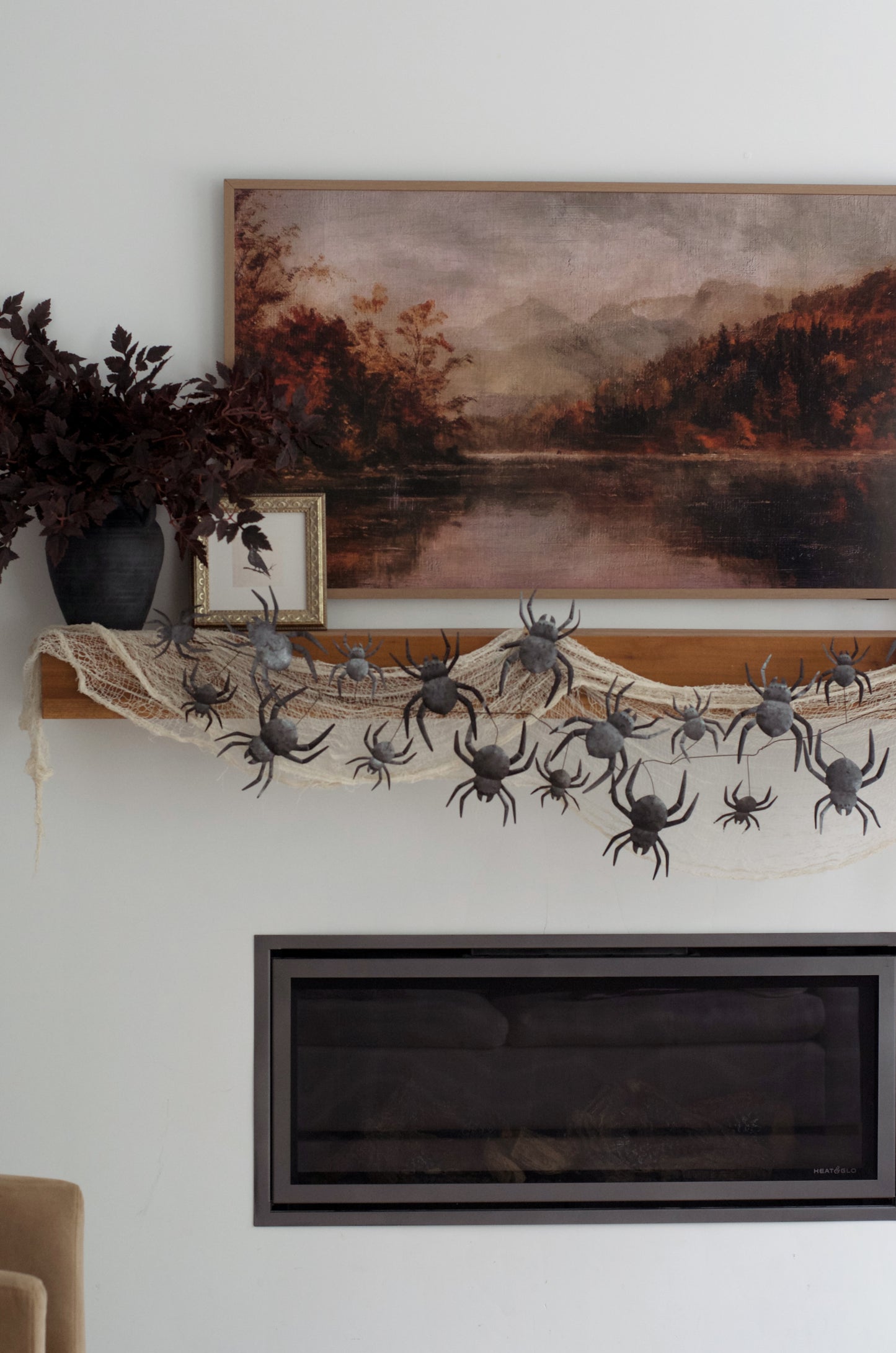 A Halloween wall decoration display featuring a vertical arrangement of black plastic spiders draped over a beige netting backdrop, next to a rustic brown and beige vertical panel, with a small mirror and dark foliage at the top.