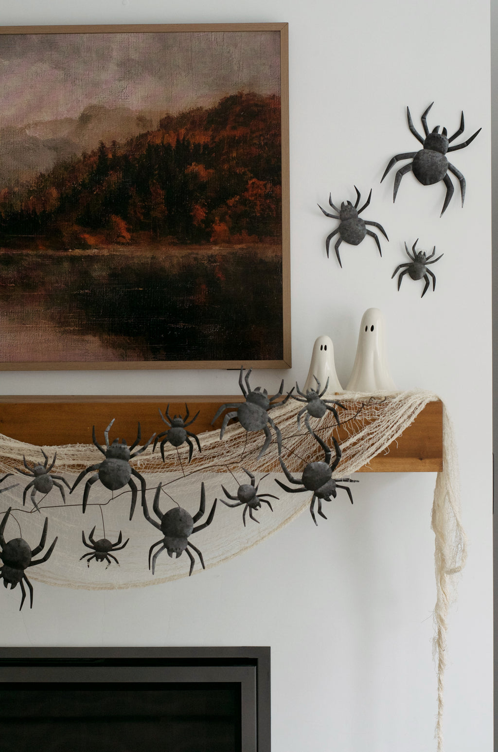 Halloween wall decor featuring black plastic toy spiders, a white spider-shaped candle holder, and a dried spiderweb hanging over a wooden ledge, styled against a white wall next to framed art.