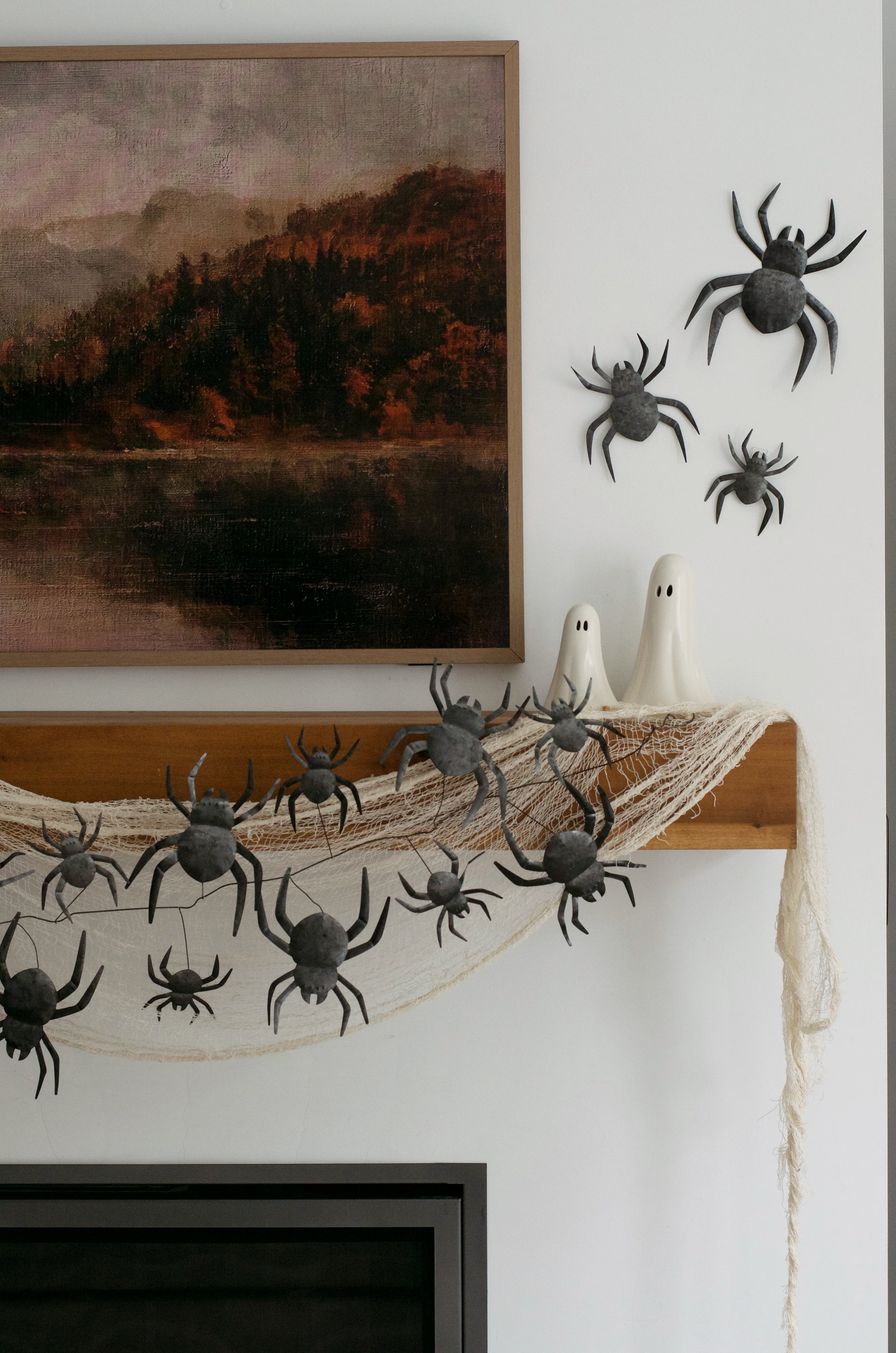 Halloween wall decor featuring black plastic toy spiders, a white spider-shaped candle holder, and a dried spiderweb hanging over a wooden ledge, styled against a white wall next to framed art.