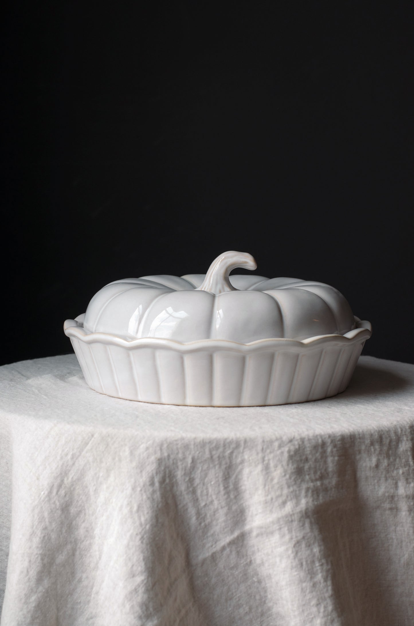 A white ceramic lidded dish or tureen shaped like a pumpkin, set on a draped white linen cloth against a dark black background.