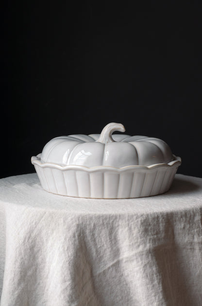 A white ceramic lidded dish or tureen shaped like a pumpkin, set on a draped white linen cloth against a dark black background.