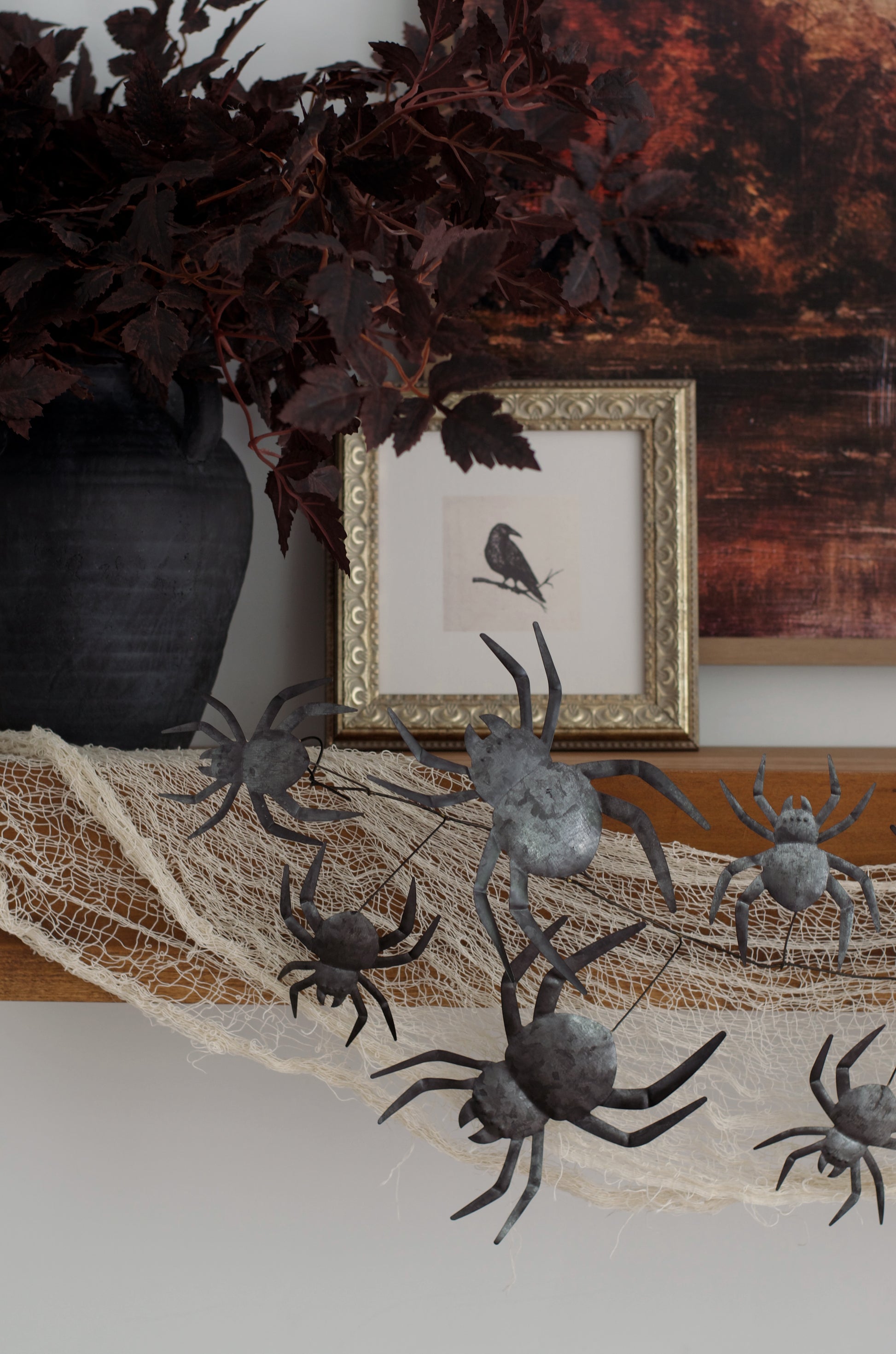 Halloween decorative metal spiders draped over a white cobweb banner on a wooden mantel, styled with a black vase, dark leaves, and a framed bird print.