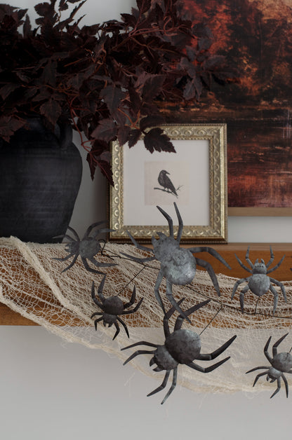 Halloween decorative metal spiders draped over a white cobweb banner on a wooden mantel, styled with a black vase, dark leaves, and a framed bird print.